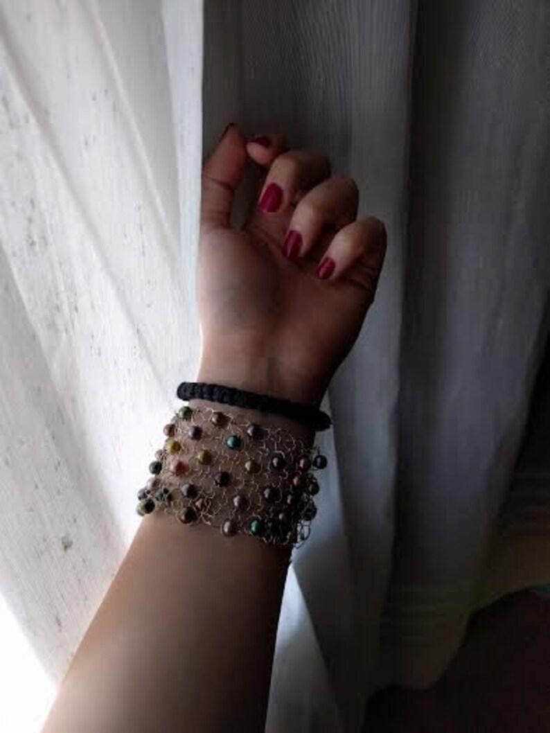May include: A wrist adorned with two bracelets. The top bracelet is a simple black band. Below, a wider bracelet features a delicate wire mesh design, embellished with iridescent beads in shades of brown, green, and gold. The background is a sheer white curtain.