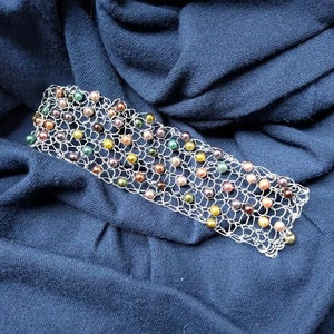 May include: A silver-toned bracelet with a delicate, openwork design. The bracelet is adorned with a variety of colorful beads, including green, yellow, brown, and purple. The bracelet is displayed on a dark blue fabric.