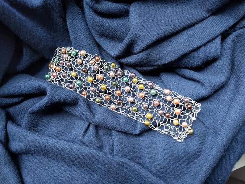 May include: A silver-toned bracelet with a delicate, openwork design. The bracelet is adorned with a variety of colorful beads, including green, yellow, brown, and purple. The bracelet is displayed on a dark blue fabric.