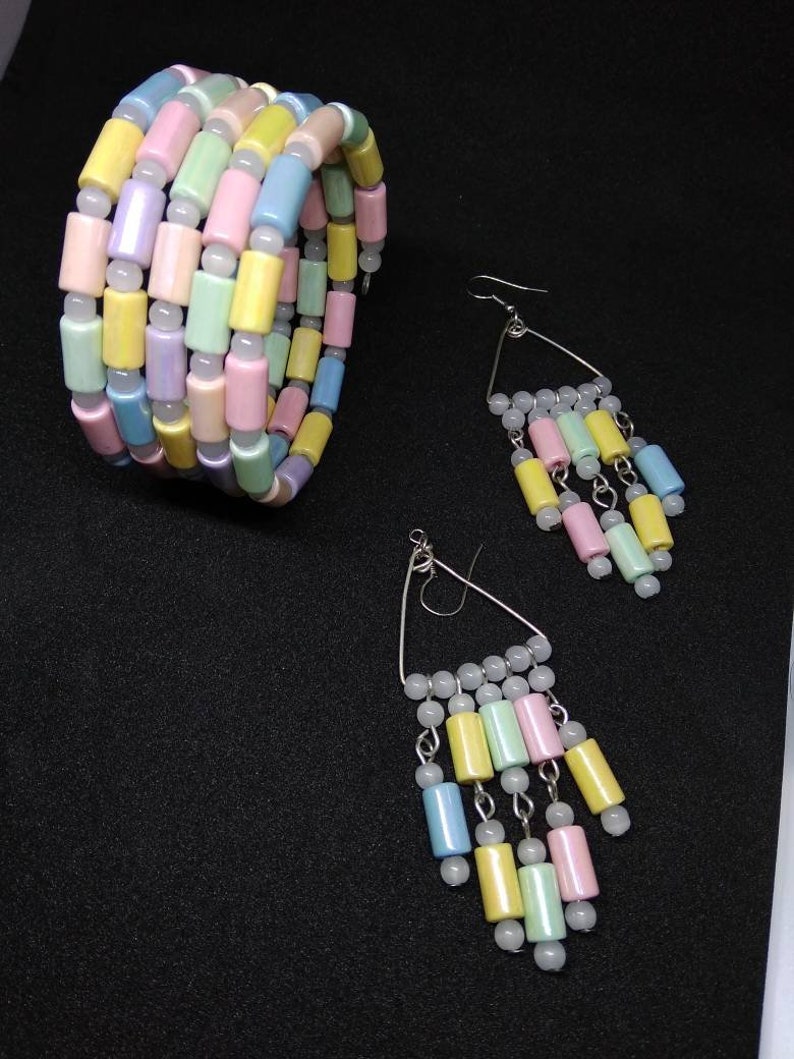 May include: A pastel-colored jewelry set featuring a bracelet and earrings. The bracelet is made of cylindrical beads in shades of pink, yellow, blue, and green. The earrings have a triangular design with dangling beads in matching colors. The jewelry is set against a black background.