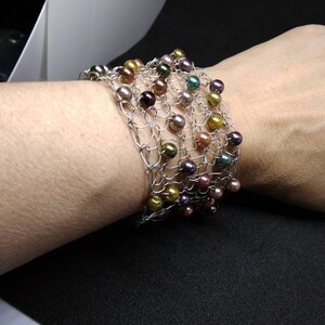 May include: A silver-toned wire bracelet featuring a woven design, adorned with a variety of colorful, round beads. The beads are in shades of green, gold, brown, and purple. The bracelet is designed to be worn around the wrist.