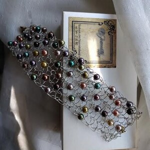 May include: A silver-toned bracelet with a woven design, adorned with multi-colored pearls. The pearls are in shades of gold, green, purple, and brown. The bracelet rests on a white surface next to a box with a label.