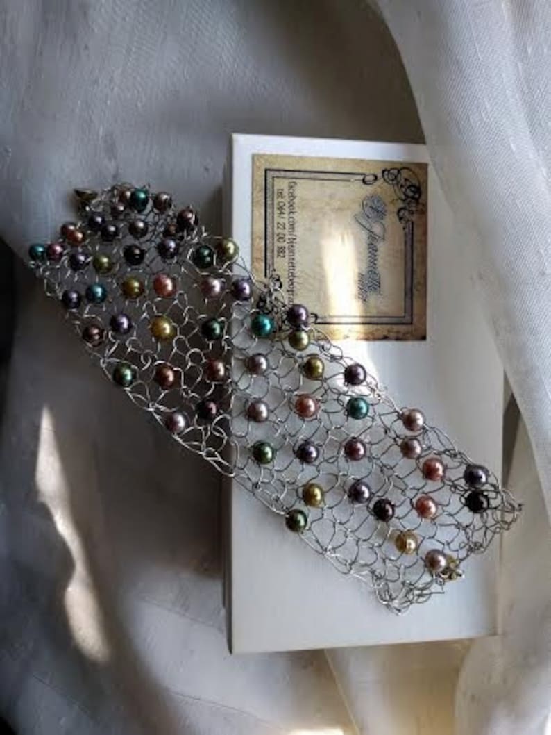 May include: A silver-toned bracelet with a woven design, adorned with multi-colored pearls. The pearls are in shades of gold, green, purple, and brown. The bracelet rests on a white surface next to a box with a label.