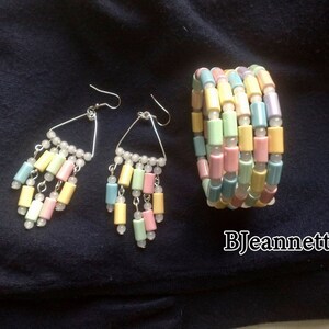 May include: Pastel-colored jewelry set featuring a bracelet and earrings. The earrings have a triangular design with dangling cylindrical beads in shades of pink, yellow, blue, and green. The bracelet is made of similar beads. The jewelry is set against a dark blue background.