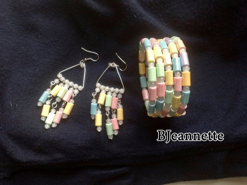 May include: Pastel-colored jewelry set featuring a bracelet and earrings. The earrings have a triangular design with dangling cylindrical beads in shades of pink, yellow, blue, and green. The bracelet is made of similar beads. The jewelry is set against a dark blue background.