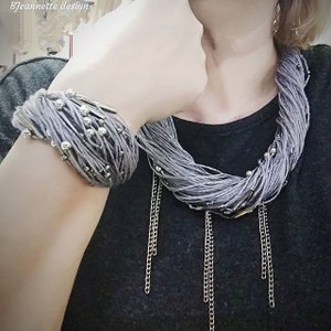 May include: A gray necklace and bracelet set. The necklace is made of multiple strands of gray thread, accented with silver beads and dangling silver chains. The bracelet has a similar design, with silver beads and a silver clasp. The jewelry is displayed on a dark background.
