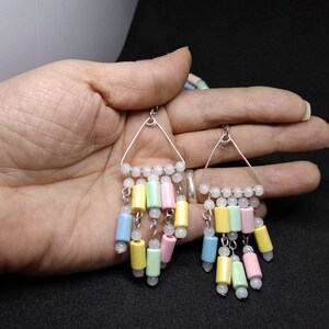 May include: A pair of pastel-colored dangle earrings. Each earring features a silver triangle frame with a row of white beads at the top. Below, cylindrical beads in yellow, green, pink, and blue hang from the frame, interspersed with white beads.