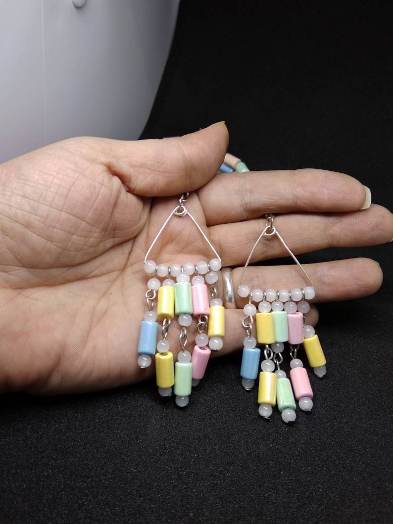 May include: A pair of pastel-colored dangle earrings. Each earring features a silver triangle frame with a row of white beads at the top. Below, cylindrical beads in yellow, green, pink, and blue hang from the frame, interspersed with white beads.