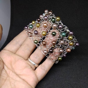 May include: A silver-toned metal bracelet with a woven design, adorned with colorful pearls in shades of green, gold, purple, and brown. The bracelet is held in a hand against a dark background.