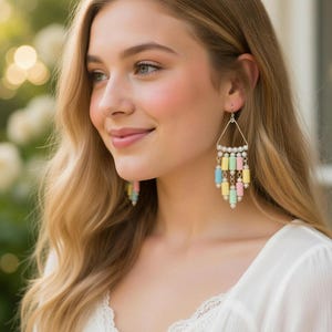 May include: A close-up of a person wearing chandelier earrings. The earrings have a silver triangle top with small white beads and pastel-colored cylindrical beads hanging below. The person has blonde hair and is wearing a white top.