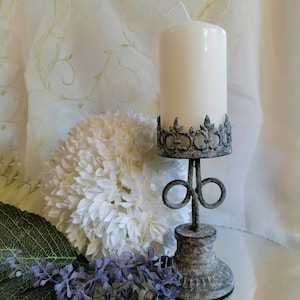 May include: A white pillar candle sits on a gray metal candle holder with a decorative design. The candle holder has a round base and a decorative top. The candle holder is sitting on a reflective surface.