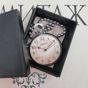 Peut inclure: Une montre de poche Raketa vintage avec une chaîne argentée, présentée dans une boîte noire. Le cadran de la montre est crème avec des chiffres noirs et rouges. Une montre classique.