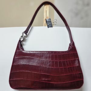 Peut inclure: Un sac à bandoulière bordeaux avec une texture peau de crocodile. Le sac a une anse incurvée et un détail de chaîne argentée sur un côté. Le sac est sur une surface blanche.