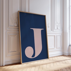 May include: A framed art print featuring a large, stylized letter "J" in a soft pink hue against a deep navy blue background. The artwork is displayed in a room with white walls and a wooden floor, leaning against the wall.