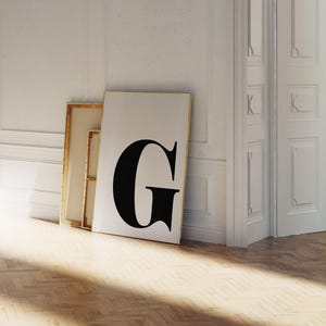 Puede incluir: Un gran cuadro enmarcado con la letra "G" negra sobre fondo blanco. La obra de arte se apoya en una pared blanca, junto a otras piezas enmarcadas, sobre un suelo de madera. La habitación es luminosa, con luz solar.