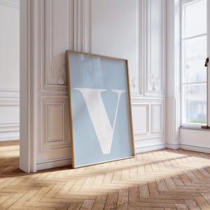 May include: A framed art print featuring a large white letter "V" on a light blue background. The print has a thin, light brown frame and is leaning against a white wall with decorative molding. The floor is made of light brown wood.