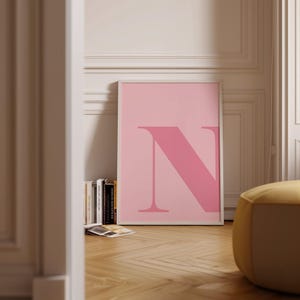May include: A framed pink art print featuring a large letter "N" in a darker shade of pink. The artwork is leaning against a wall, with a stack of books and a yellow ottoman in the foreground. The room has a light pink and white color scheme.
