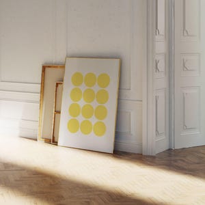 May include: A framed art print featuring a pattern of twelve yellow circles on a white background. The artwork leans against a white wall, next to a framed canvas with a gold frame. The room has a wooden floor and white doors.