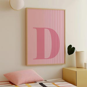 Pode incluir: Uma arte de parede rosa emoldurada com a letra "D" em um tom de rosa mais escuro. O fundo tem listras verticais. A moldura é de cor de madeira clara. A arte está pendurada acima de uma cama com uma almofada rosa.