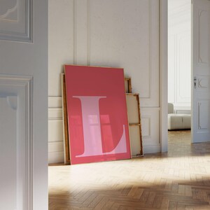 May include: A large, framed pink art print with a light pink letter "L" on a solid pink background. The artwork leans against a white wall, with other framed pieces behind it. The room has a hardwood floor and an open doorway.