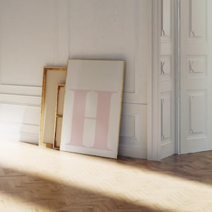 Peut inclure: Une grande affiche rectangulaire avec la lettre "H" rose pâle sur fond blanc. L'affiche est appuyée contre un mur, à côté d'une toile encadrée avec un bord doré. La pièce a un parquet et des murs blancs.