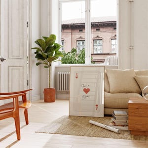 May include: A framed art print featuring a playing card design with the text "Take a Chance" and a red heart. The print is leaning against a beige sofa in a well-lit room with a large window and a potted plant.