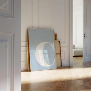 May include: A large framed print featuring a light blue background with a white letter "C". The print is leaning against a wall in a room with a wooden floor. The frame has a gold border.