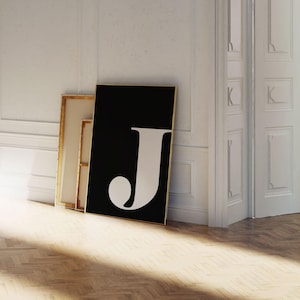 Può includere: Una stampa artistica incorniciata in bianco e nero con una grande lettera "J" bianca. L'opera d'arte è appoggiata a una parete bianca e a un telaio di una porta, con una cornice dorata. Il pavimento è in legno.