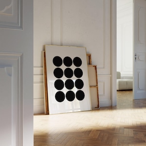 May include: A large framed art print featuring a minimalist design of twelve black circles arranged in a grid pattern on a white background. The artwork leans against a wall in a room with hardwood floors and white walls.