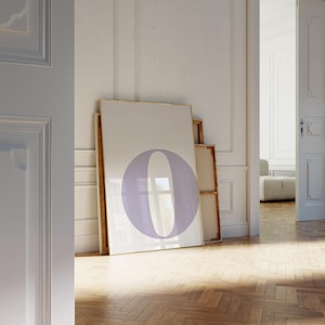 以下が含まれることがあります： 白い背景にラベンダー色の文字「O」が描かれた、大きな額入りのプリント。木製の床と白い壁のある部屋の壁に立てかけられています。フレームは金色のトリムです。
