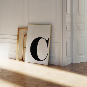 May include: A large framed print featuring a bold black letter "C" against a white background. The artwork leans against a white wall, with a gold frame. The image is set in a room with a wooden floor and a white door.