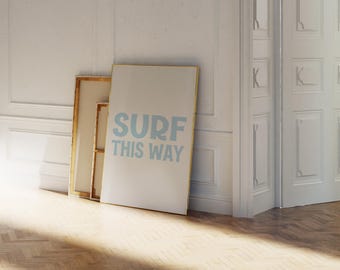 Puede incluir: Tres obras de arte enmarcadas apoyadas contra una pared blanca. La pieza más grande tiene las palabras "SURF THIS WAY" en azul claro. Los marcos tienen tonos dorados y de madera natural. El suelo es de madera clara.