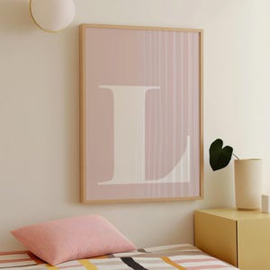 May include: A framed art print featuring a large white letter "L" against a soft pink background with vertical lines. The artwork is encased in a light wood frame, creating a minimalist aesthetic. The print is displayed on a wall above a bed.