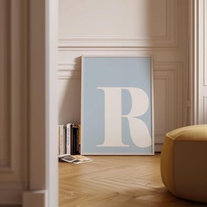 May include: A framed poster featuring a large white letter "R" against a light blue background. The poster is leaning against a wall, with a stack of books and a yellow ottoman in the foreground. The room has a hardwood floor.