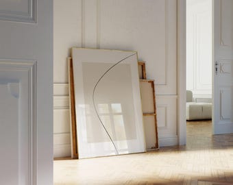 May include: A large framed artwork with a gold border leans against a white wall. The artwork features a beige background with a black curved line. Other framed pieces are stacked behind it. The room has a hardwood floor and an open doorway.