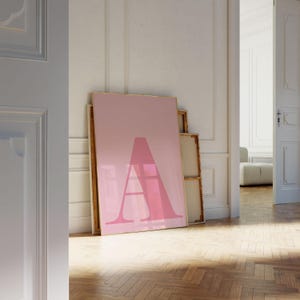 Puede incluir: Una gran impresión artística enmarcada en rosa con la letra "A" estilizada en un tono rosa más claro. La obra de arte se apoya contra una pared en una habitación con suelo de madera y paredes blancas. La impresión es parte de un conjunto de impresiones artísticas.