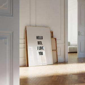 Puede incluir: Una impresión artística enmarcada con el texto "HELLO BED, I LOVE YOU" en negro sobre un fondo blanco. La impresión está apoyada contra una pared en una habitación con suelo de parquet y una puerta.
