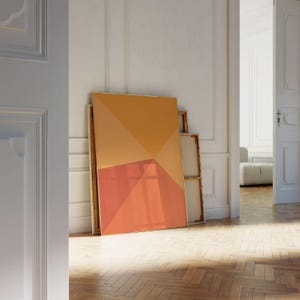 May include: An abstract geometric painting with a gold frame, featuring orange and coral triangular shapes. The artwork leans against a white wall in a room with a wooden floor. Additional framed canvases are visible.