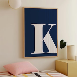 Puede incluir: Un cuadro enmarcado con una gran letra "K" blanca sobre un fondo azul marino. La obra de arte está en un marco de madera y cuelga de una pared beige. La habitación también incluye un cojín rosa y una mesita de noche amarilla.