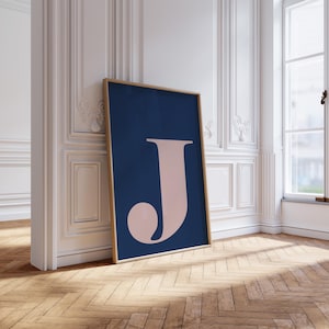 May include: A framed art print featuring a large, stylized letter "J" in a soft pink hue against a deep navy blue background. The artwork is displayed in a room with light-colored walls and a wooden floor, leaning against the wall.
