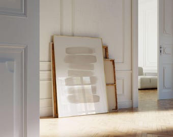 Puede incluir: Una gran impresión artística abstracta enmarcada con pinceladas horizontales en tonos beige y gris. La obra de arte se apoya contra una pared blanca, con otras piezas enmarcadas visibles. La habitación tiene un suelo de madera y una puerta que conduce a otra habitación.