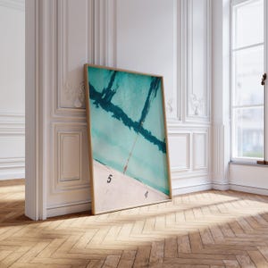 May include: A framed art print depicting a turquoise pool scene with dark blue lines and a red rope. The bottom of the print shows the numbers 4 and 5. The frame is light brown. The artwork is leaning against a white wall.