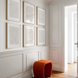May include: Six framed abstract art prints with thin black lines on a beige background, mounted on a white wall. The frames are light brown. An orange curved stool sits below the artwork. The room has white paneling and a doorway.