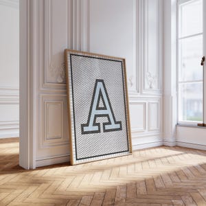 Peut inclure: Une affiche encadrée avec la lettre "A" en bleu clair. La lettre est sur un fond blanc avec un motif de grille noire. Le cadre est de couleur marron clair. L'œuvre est appuyée contre un mur blanc. Le sol est en bois.