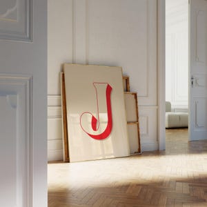 May include: A large framed print featuring the letter "J" in a bold, red, serif font. The letter has a shadow effect, adding depth. The artwork leans against a white wall in a room with a wooden floor.