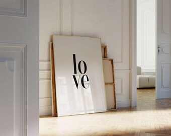 May include: A large white framed art print with the word "love" in black, leaning against a wall. The print has a gold frame. Other framed art pieces are behind it. The room has white walls and a wooden floor.