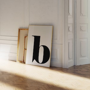 May include: A large framed art print featuring a bold black letter "b" against a white background. The print leans against a wall, next to a smaller, unframed canvas. The room has a hardwood floor and white walls.