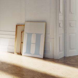 May include: A large canvas print featuring a light blue letter "H" against a cream background. The artwork leans against a white wall, next to a framed canvas. The room has a hardwood floor and white trim.