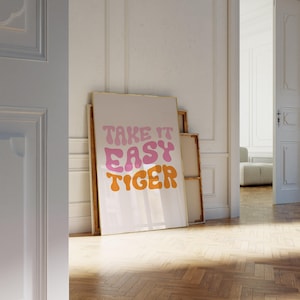 May include: A framed art print with the words "TAKE IT EASY TIGER" in pink and orange retro font. The artwork leans against a white wall in a room with a wooden floor. The print has a gold frame.