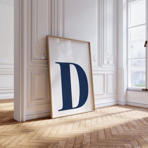 Può includere: Una stampa incorniciata con la lettera "D" blu navy su sfondo bianco. L'opera è in una cornice di legno marrone chiaro, appoggiata a una parete bianca. La stanza ha un pavimento in legno e una finestra.
